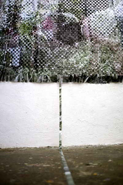 Zwei große Farbfotos wurden in streifen geschnitten und kreuzweise miteinander verwoben. Das resultierende Objekt hängt an einer weißen Wand. Ein einziger Streifen eines der beides Fotos reicht bis auf den Boden, und kommt der Kamera entgegen.