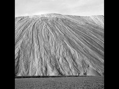 Schwarzweißfoto einer Landschaft. Hinter einem Feld hebt sich eine riesige Abraumhalde aus Salz. Die Oberfläche ist von Rillen und trockenen Flüssen überzogen. Oben ist ein schmaler Streifen bewölkter Himmel zu sehen.