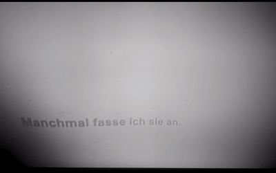 Dunkelgraue Schrift verzerrt auf hellgrauem Hintergrund: Manchmal fasse ich sie an.